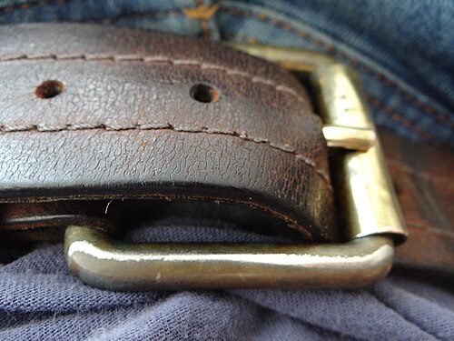 Close-up of a person's waist showing a brass rectangular belt buckle with a single prong on a brown leather belt worn over blue jeans with a t-shirt.