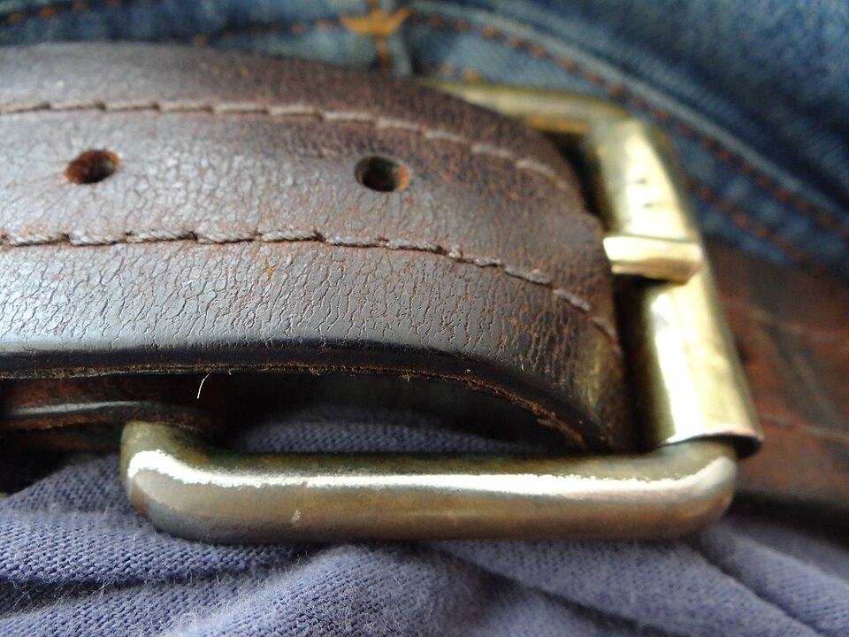 Close-up of a person's waist showing a brass rectangular belt buckle with a single prong on a brown leather belt worn over blue jeans with a t-shirt.