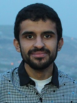 Head-and-shoulders photo of a young man with short dark hair and a short beard, smiling slightly, wearing a black-and-white checked shirt