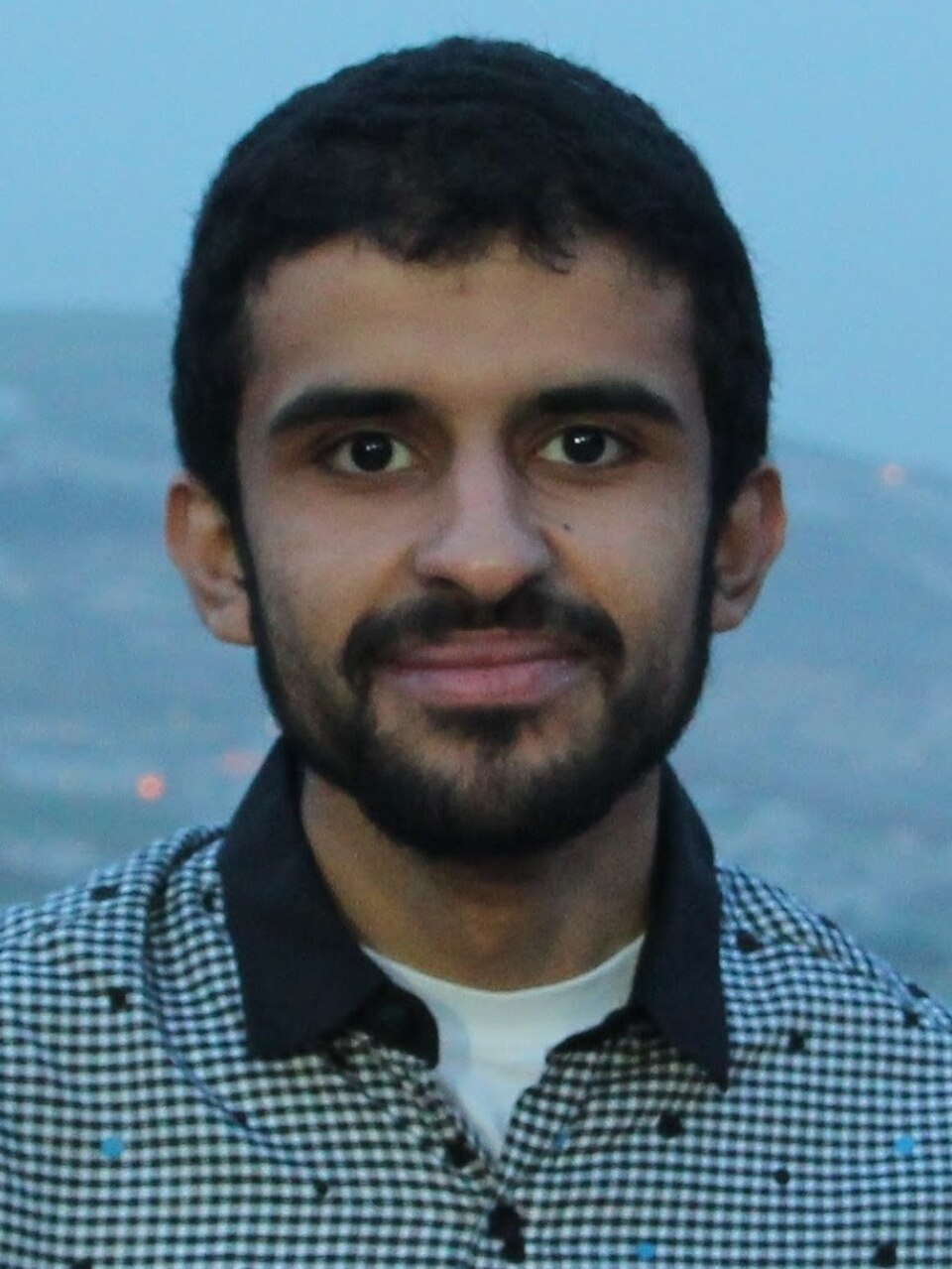 Head-and-shoulders photo of a young man with short dark hair and a short beard, smiling slightly, wearing a black-and-white checked shirt with a dark collar, outdoors at dusk.